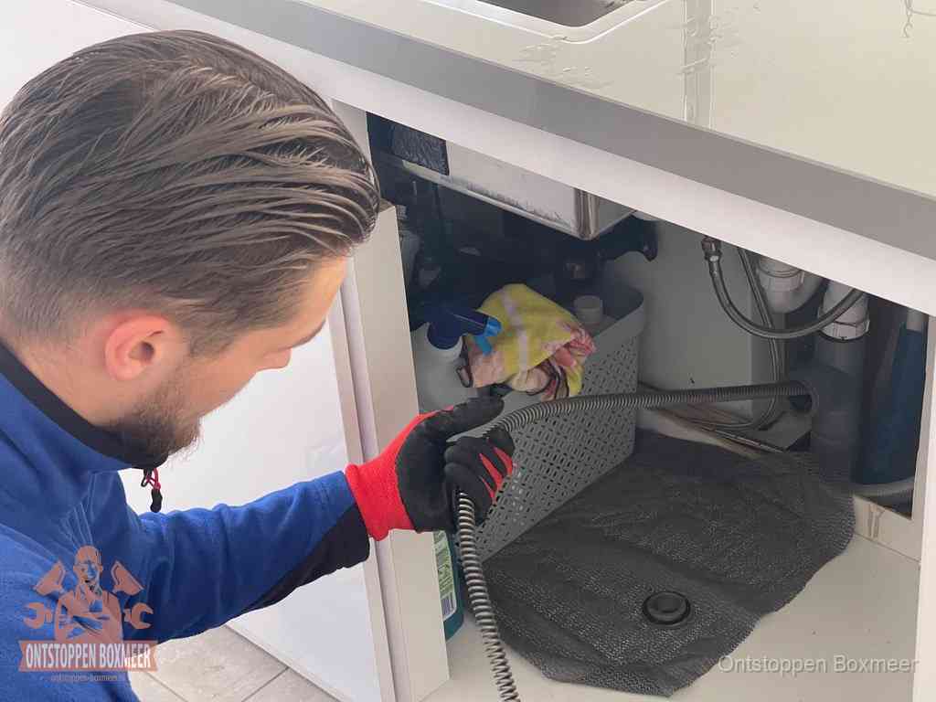 Een monteur voert een flexibele ontstoppingsveer in onder het keukenkastje, langs de sifon, om de gootsteen te ontstoppen in Boxmeer Een monteur voert een flexibele ontstoppingsveer in onder het keukenkastje, langs de sifon, om de gootsteen te ontstoppen in Boxmeer