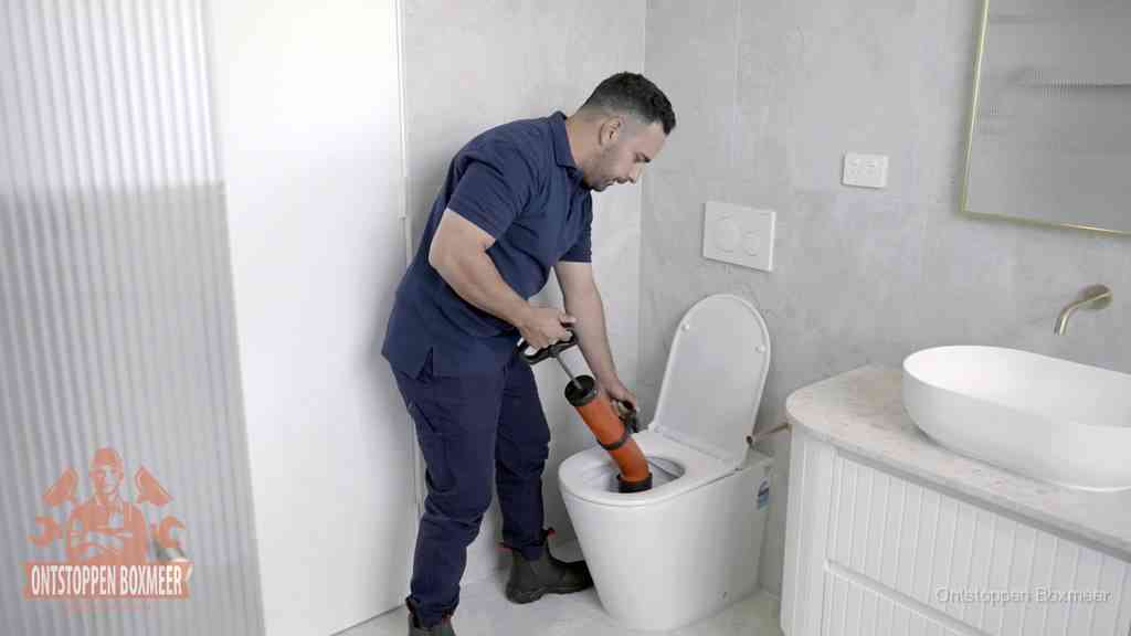 Een monteur buigt over een wandcloset en duwt met de hand een persluchtontstopper in de toiletpot om de verstopping handmatig los te maken in Boxmeer Een monteur buigt over een wandcloset en duwt met de hand een persluchtontstopper in de toiletpot om de verstopping handmatig los te maken in Boxmeer