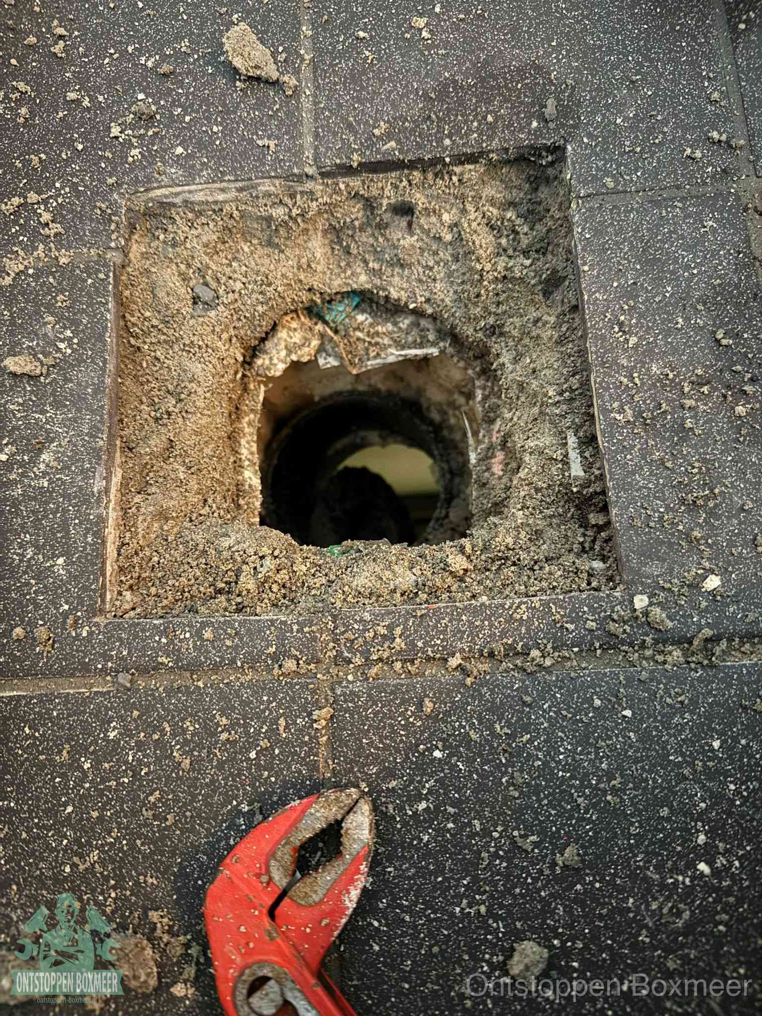 Een geopend rioolputje in een betontegelvloer met losgemaakt rooster en een zichtbare ronde afvoerbuis, het achterhalen waar de afvoerverstopping zit in Boxmeer