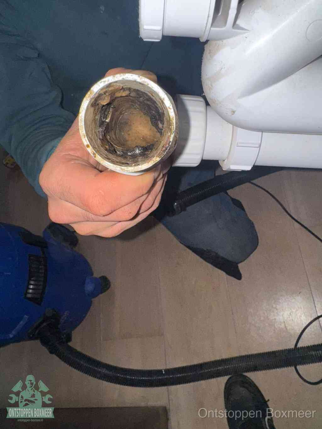 Een hand houdt een losgekoppeld PVC-afvoerbuisstuk vast dat van binnen volledig is dichtgeslibd met vet, vuil en aanslag, de meest voorkomende oorzaak van een verstopte afvoer in Boxmeer