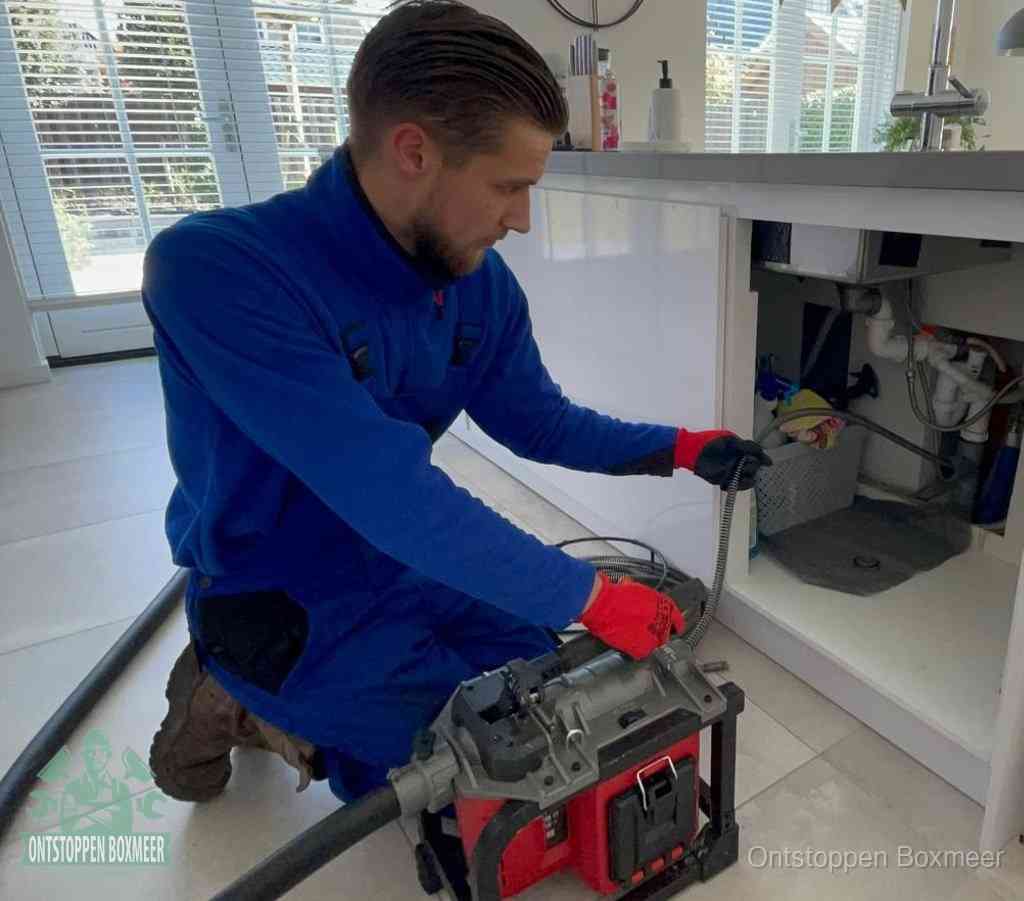 Een monteur met rode handschoenen knielt in een keuken en voert een elektrische ontstoppingsveer in tijdens het professioneel afvoer ontstoppen in Boxmeer